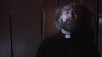 Movie still from “The Amityville Moon” (2021), directed by Thomas J. Churchill – A man with a beard wearing a priest's outfit; Close Up shot, Low angle