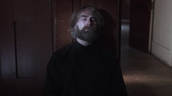 Movie still from “The Amityville Moon” (2021), directed by Thomas J. Churchill – A man with a long beard is standing in a dark room; Medium shot, Low angle