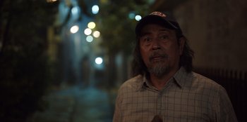 Movie still from “The Big 4” (2022), directed by Timo Tjahjanto – An older man wearing a baseball cap standing on a street; Close Up shot, Over the shoulder angle