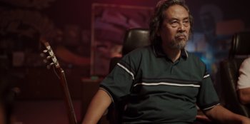 Movie still from “The Big 4” (2022), directed by Timo Tjahjanto – A man sitting in a chair in front of a painting; Medium shot, Over the shoulder angle