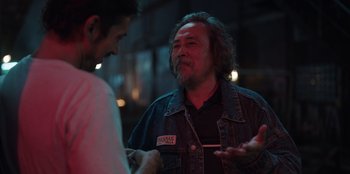 Movie still from “The Big 4” (2022), directed by Timo Tjahjanto – A man with long hair talking to another man in a dark room; Medium shot, Over the shoulder angle