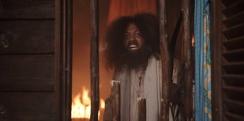 Movie still from “The Big 4” (2022), directed by Timo Tjahjanto – A man with long hair and a beard is standing in front of a fire; Medium shot, Over the shoulder angle