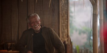 Movie still from “The Big 4” (2022), directed by Timo Tjahjanto – An older man smiling while sitting on a chair; Medium shot, Low angle