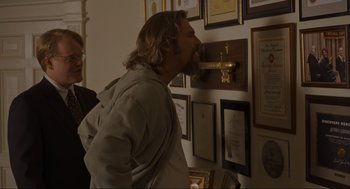 Movie still from “The Big Lebowski” (1998), directed by Ethan Coen – A man with long hair and a long beard standing in front of a wall with many awards on it; Medium shot, Over the shoulder angle