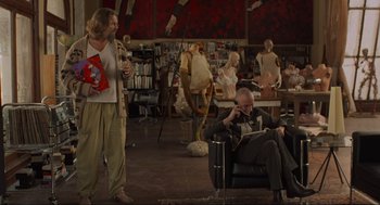 Movie still from “The Big Lebowski” (1998), directed by Ethan Coen – A man sitting on a chair in a room with other people; Wide shot, Low angle