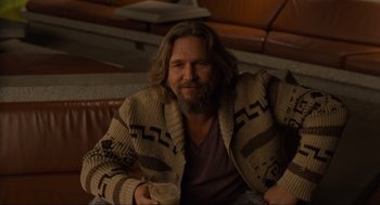 Movie still from “The Big Lebowski” (1998), directed by Ethan Coen – A man sitting on top of a couch holding a drink; Medium shot, Over the shoulder angle