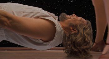 Movie still from “The Big Lebowski” (1998), directed by Ethan Coen – A man with long hair and a beard is doing a yoga pose; Medium shot, Overhead angle