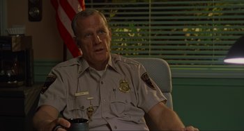 Movie still from “The Big Lebowski” (1998), directed by Ethan Coen – A police officer sitting in a chair holding a coffee mug; Close Up shot, Over the shoulder angle