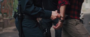 Movie still from “The Binge” (2020), directed by Jeremy Garelick – A police officer holding a pair of handcuffed hands; Medium shot, Low angle