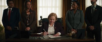 Movie still from “The Binge” (2020), directed by Jeremy Garelick – A woman sitting at a desk writing on a piece of paper; Medium shot, Low angle