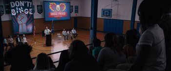 Movie still from “The Binge” (2020), directed by Jeremy Garelick – A group of people sitting in front of an audience in a gymnasium; Extreme Wide shot, Over the shoulder angle