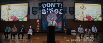 Movie still from “The Binge” (2020), directed by Jeremy Garelick – A man standing at a podium in front of an american flag; Extreme Wide shot, Over the shoulder angle