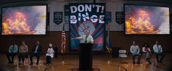 Movie still from “The Binge” (2020), directed by Jeremy Garelick – A man standing at a podium in front of an american flag; Extreme Wide shot, Over the shoulder angle