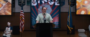 Movie still from “The Binge” (2020), directed by Jeremy Garelick – A man standing at a podium in front of a poster; Medium shot, Over the shoulder angle