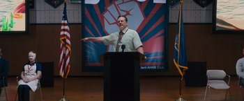 Movie still from “The Binge” (2020), directed by Jeremy Garelick – A man standing at a podium in front of a poster; Medium shot, Low angle