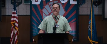 Movie still from “The Binge” (2020), directed by Jeremy Garelick – A man standing at a podium in front of a microphone; Medium shot, Low angle