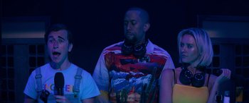 Movie still from “The Binge” (2020), directed by Jeremy Garelick – A man with headphones on standing next to another man; Medium shot, Over the shoulder angle