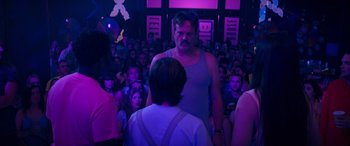 Movie still from “The Binge” (2020), directed by Jeremy Garelick – A crowd of people sitting in front of a man in a gray tank top; Medium shot, Over the shoulder angle