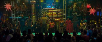 Movie still from “The Binge” (2020), directed by Jeremy Garelick – A group of people standing on a stage in front of an american flag; Extreme Wide shot, Low angle