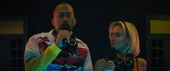 Movie still from “The Binge” (2020), directed by Jeremy Garelick – A man and a woman holding microphones in a dark room; Close Up shot, Low angle