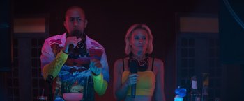 Movie still from “The Binge” (2020), directed by Jeremy Garelick – A man and a woman standing next to each other holding microphones; Medium shot, Low angle