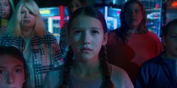 Movie still from “It's A Wonderful Binge” (2022), directed by Jordan VanDina – A young girl with plaited pigtails in front of a group of people; Close Up shot, Over the shoulder angle