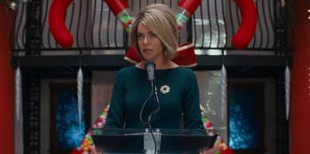 Movie still from “It's A Wonderful Binge” (2022), directed by Jordan VanDina – A woman standing at a podium speaking into a microphone; Medium shot, Low angle
