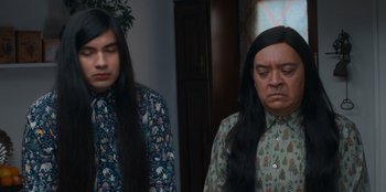 Movie still from “It's A Wonderful Binge” (2022), directed by Jordan VanDina – A man with long hair is standing next to another man with long black hair; Medium shot, Over the shoulder angle