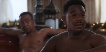 Movie still from “It's A Wonderful Binge” (2022), directed by Jordan VanDina – Two shirtless men sitting in a living room; Close Up shot, Over the shoulder angle