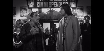 Movie still from “It's A Wonderful Binge” (2022), directed by Jordan VanDina – Two people standing next to each other in front of a vote for mayor sign; Medium shot, Over the shoulder angle