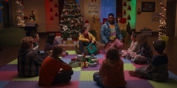 Movie still from “It's A Wonderful Binge” (2022), directed by Jordan VanDina – A group of children sitting on the ground in front of a christmas tree; Wide shot, Over the shoulder angle