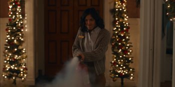 Movie still from “It's A Wonderful Binge” (2022), directed by Jordan VanDina – A woman holding a fire extinguisher in front of a christmas tree; Medium shot, Over the shoulder angle