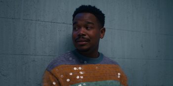 Movie still from “It's A Wonderful Binge” (2022), directed by Jordan VanDina – A man in a colorful sweater is standing in front of a brick wall; Close Up shot, Low angle