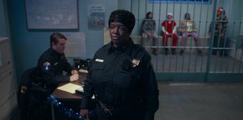 Movie still from “It's A Wonderful Binge” (2022), directed by Jordan VanDina – A woman in a police uniform standing in front of a jail cell; Medium shot, Over the shoulder angle