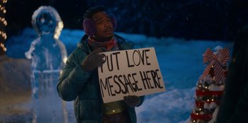 Movie still from “It's A Wonderful Binge” (2022), directed by Jordan VanDina – A man holding up a sign that says " put love message here "; Medium shot, Low angle