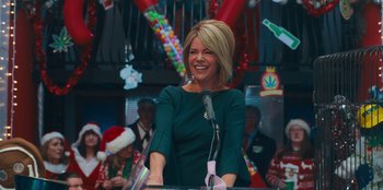 Movie still from “It's A Wonderful Binge” (2022), directed by Jordan VanDina – A woman standing at a podium in front of a crowd; Medium shot, Over the shoulder angle