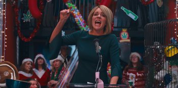 Movie still from “It's A Wonderful Binge” (2022), directed by Jordan VanDina – A woman in a green dress is speaking at a podium; Medium shot, Low angle
