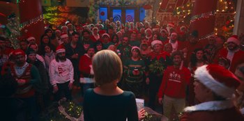 Movie still from “It's A Wonderful Binge” (2022), directed by Jordan VanDina – A group of people standing in front of a woman in a room; Wide shot, High angle