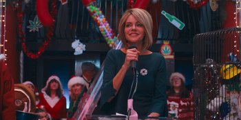 Movie still from “It's A Wonderful Binge” (2022), directed by Jordan VanDina – A woman standing at a microphone in front of a group of people; Medium shot, Over the shoulder angle