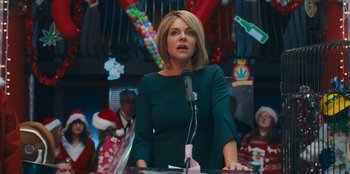 Movie still from “It's A Wonderful Binge” (2022), directed by Jordan VanDina – A woman standing at a podium speaking to a crowd; Medium shot, Over the shoulder angle