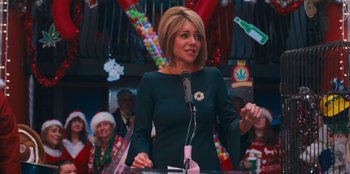 Movie still from “It's A Wonderful Binge” (2022), directed by Jordan VanDina – A woman standing at a podium in front of a crowd; Medium shot, Over the shoulder angle