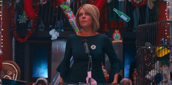 Movie still from “It's A Wonderful Binge” (2022), directed by Jordan VanDina – A woman standing at a podium in front of a microphone; Medium shot, Low angle