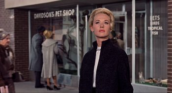 Movie still from “The Birds” (1963), directed by Alfred Hitchcock – A woman standing in front of a store with people in the background; Medium shot, Low angle