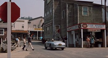 Movie still from “The Birds” (1963), directed by Alfred Hitchcock – An old car driving down a street past a building; Wide shot, High angle