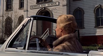 Movie still from “The Birds” (1963), directed by Alfred Hitchcock – A woman driving a car in front of a building; Medium shot, Low angle