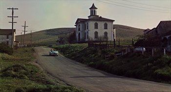 Movie still from “The Birds” (1963), directed by Alfred Hitchcock – A car driving down a road near a church; Extreme Wide shot, Low angle