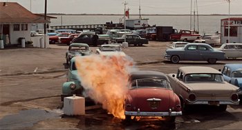 Movie still from “The Birds” (1963), directed by Alfred Hitchcock – An old car is on fire in a parking lot; Extreme Wide shot, High angle
