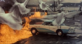 Movie still from “The Birds” (1963), directed by Alfred Hitchcock – A car that is on fire with birds flying around it; Wide shot, High angle