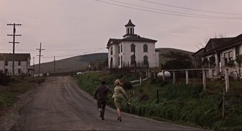 Movie still from “The Birds” (1963), directed by Alfred Hitchcock – Two people running down a dirt road near a church; Extreme Wide shot, Low angle