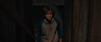 Movie still from “The Black Phone” (2021), directed by Scott Derrickson – A young boy standing in a dark room wearing a blue and gray shirt; Medium shot, Over the shoulder angle